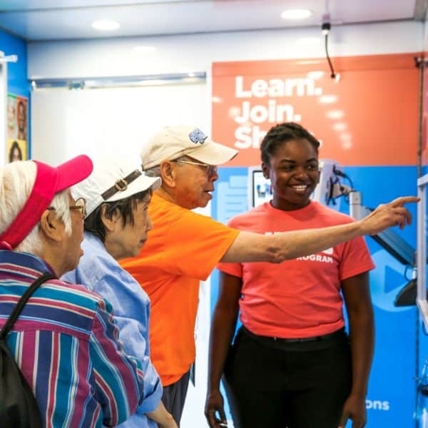 Three elderly Asian people wearing hats look at an exhibit in the All of Us Journey, with one pointing at something he sees on the wall. A young Black woman smiles and looks at where he is pointing.