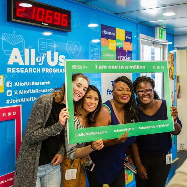 A racially diverse group of four women pose inside a jumbo photo frame on the All of Us Journey.
