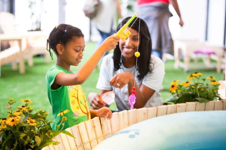 Young Black girl in green shirt plays a fishing game while being supervised by a dark haired woman.